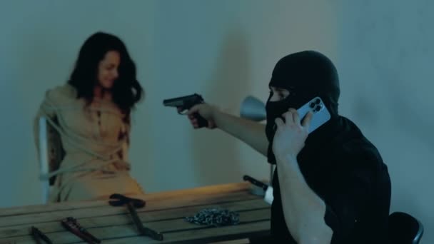 Férfi emberrabló mobilon beszél és megfenyeget egy nőt egy elhagyatott épületben. Fekete maszkos férfi fegyverrel. Emberrablás elmélet. Valós idejű videó. - Felvétel, videó