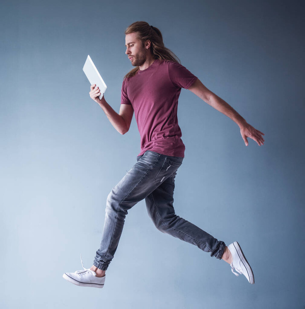 Elegáns fiatalember vállig érő szőke haja és alkalmi ruhák gazdaság egy digitális tabletta, míg jumping, szürke háttér - Fotó, kép