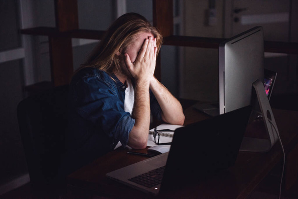 Knappe jonge zakenman blond haar bedekt met schouder lengte zijn gezicht moe of werken met een computer bij nacht - Foto, afbeelding