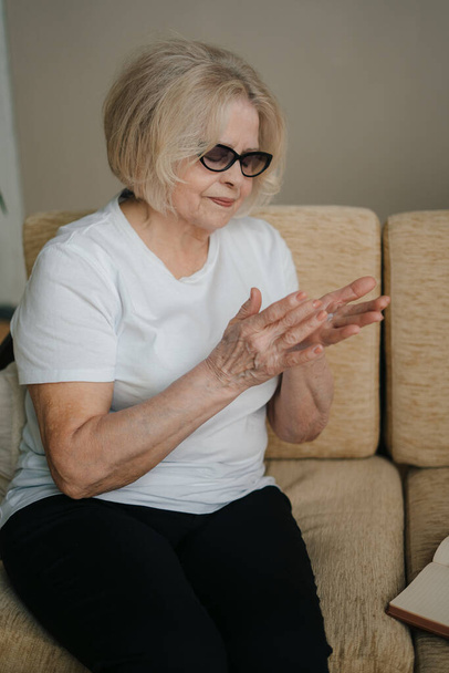 Старша жінка з коротким сірим волоссям наносить крем на руки, насолоджуючись новими косметичними засобами, що сидять на бежевому дивані вдома. Догляд за шкірою, рутина вдома і користь - Фото, зображення