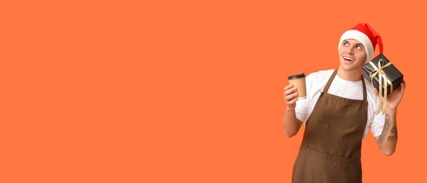 Male barista with Christmas gift and cup of coffee on orange background with space for text - Photo, image