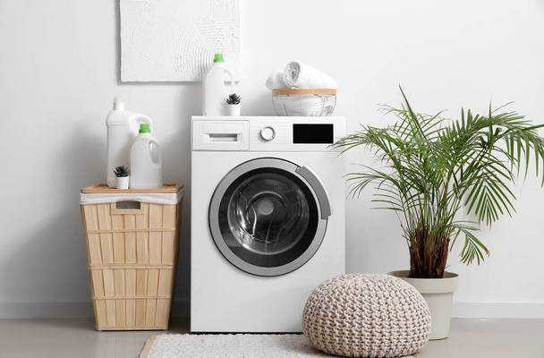 Máquina de lavar com garrafas de detergente, cesta de roupa e pufe perto da parede branca - Foto, Imagem