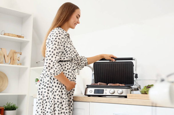 Bela jovem grávida cozinhando salsichas na moderna churrasqueira elétrica na cozinha - Foto, Imagem