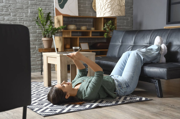Mladá žena pomocí mobilního telefonu na koberci v obývacím pokoji - Fotografie, Obrázek