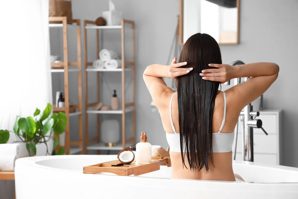 Belle jeune femme appliquant de l'huile de coco sur ses cheveux dans la salle de bain, vue arrière - Photo, image