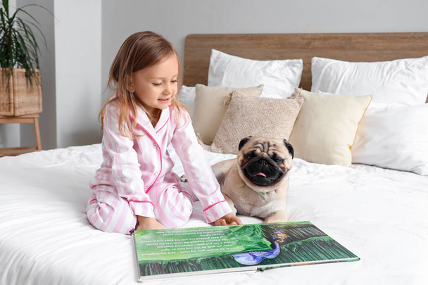 Маленькая девочка с милой собакой-мопсом и книгой в спальне - Фото, изображение