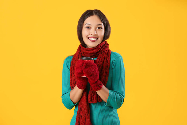 Beautiful young Asian woman in warm scarf with cup of coffee on yellow background - Photo, Image