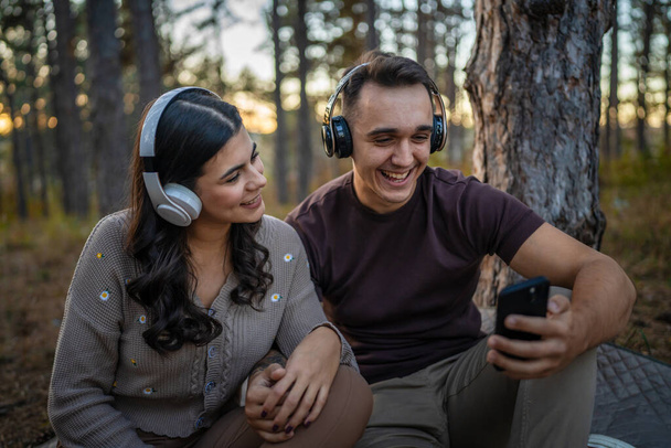 Homem e mulher jovem casal adulto na natureza ouvir música em fones de ouvido usar smartphone telefone celular ou fazer um aplicativo de chamada de vídeo - Foto, Imagem