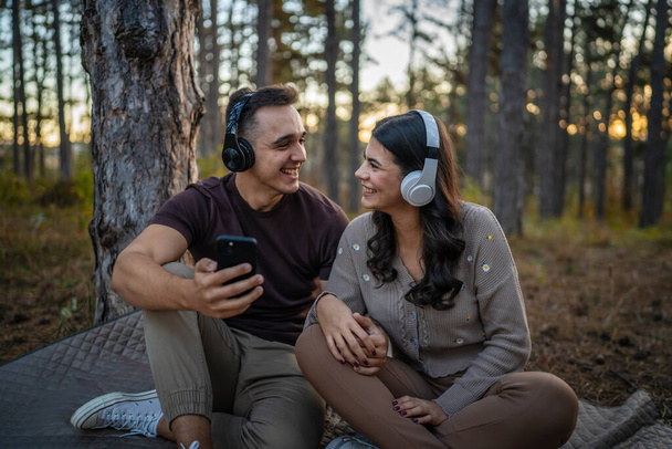 Man en vrouw jong volwassen stel in de natuur luisteren muziek op koptelefoon gebruik maken van mobiele telefoon smartphone of maak een video call app - Foto, afbeelding