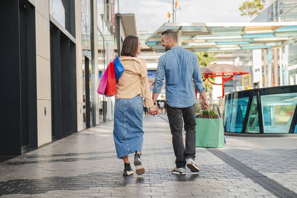 Πίσω όψη του νεαρού ζευγαριού ψώνια σε ένα εμπορικό κέντρο, μεταφέρουν χάρτινες σακούλες στα χέρια τους και περπατάνε μαζί κοιτάζοντας ο ένας τον άλλον και χαμογελώντας. Σύζυγος και γυναίκα αξιοθέατα κρατώντας τα χέρια απολαμβάνοντας σε μια ημερομηνία - Φωτογραφία, εικόνα