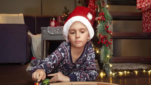 Iloinen pikkupoika pyjamassa leikkii uudella lelujunallaan ja rautatiellään, jonka hän sai joululahjaksi. Perhejuhlat talvilomilla. - Materiaali, video