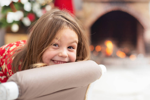 petite fille heureuse sourit doucement et drôle, repose confortablement sur une chaise sur le fond de feu dans la cheminée. Pour votre projet de vente et de publicité d'accessoires d'hiver chauds pour la cheminée. - Photo, image