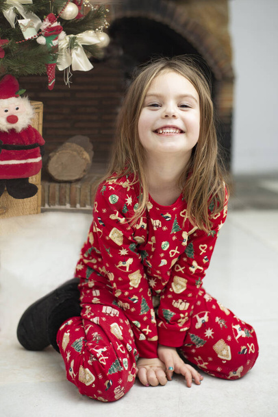 Dziewczynka w zimowej piżamie świątecznej uśmiecha się uroczo, pozując przy choince i kominku. prezentacja ciepłych zimowych ubrań i butów dziecięcych przez niewielką modelkę. Modele dla dzieci. - Zdjęcie, obraz
