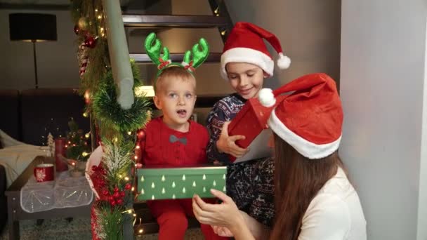 Dos chicos lindos en pijama recibieron regalos de Navidad y regalos de Santa en Año Nuevo sentados en escaleras de madera en casa con madre. Celebraciones familiares en las vacaciones de invierno - Metraje, vídeo