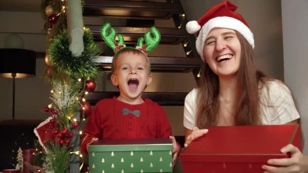 Iloinen naurava lapsi poika, jolla on äiti, jolla on joululahjoja ja esittää joulupukin laatikoita ja hymyilee kameran edessä. Perhejuhlat talvilomilla - Materiaali, video