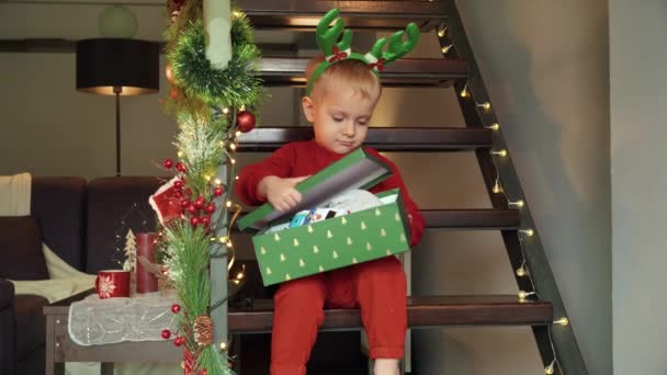 Lindo niño abre la caja de regalo de Navidad y se emociona de sus nuevos juguetes. Celebraciones familiares en las vacaciones de invierno - Imágenes, Vídeo