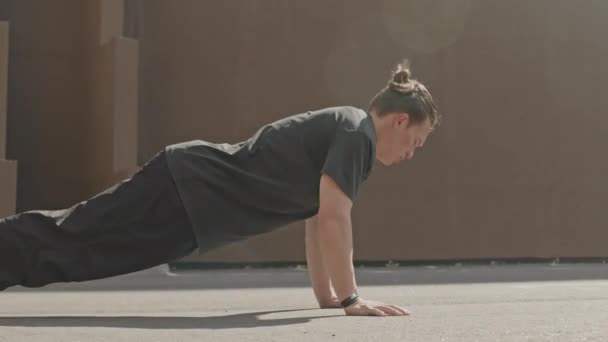 Hitaasti urheilullinen nuori valkoihoinen kaveri tekee punnerruksia ulkona auringonvalolla asfaltilla kaupunkiympäristössä - Materiaali, video