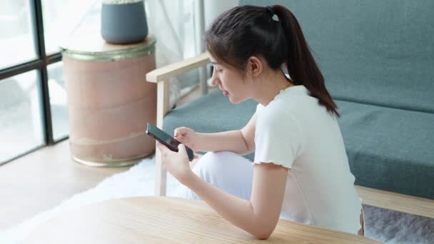 Mujer asiática relajada usando teléfonos inteligentes navegando por las redes sociales, revisando noticias, jugando juegos móviles o mensajes de texto sentados en el sofá. Milenial dama pasar tiempo en casa con tecnología gadget celular - Imágenes, Vídeo