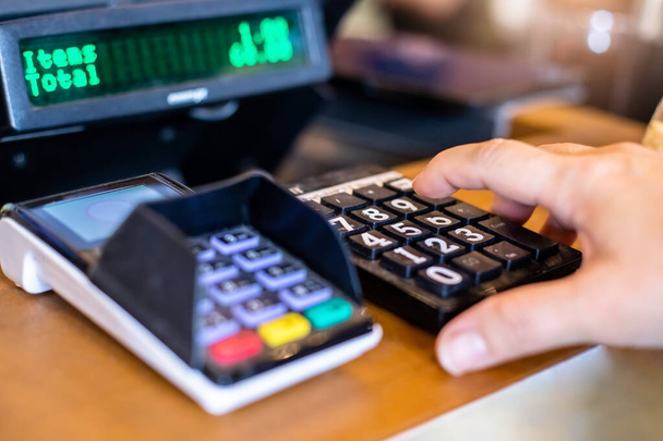 Selective focus to finger of women inserting password or pin code login on machine keypad of payment terminal. Cash desk with  payment terminal and blurry information screen in cafe. - Photo, Image