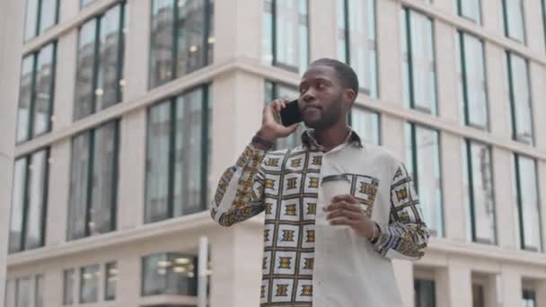 Düşük açılı orta açılı bir fotoğraf, elinde kahve fincanı olan bir genç adam akıllı telefondan konuşuyor ve etrafa bakıyor, arka planda çağdaş ofis binasına karşı duruyor. - Video, Çekim
