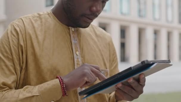 Талия молодого чернокожего человека, использующего цифровой планшет на открытом воздухе в центре города, работающего вдали от офиса - Кадры, видео