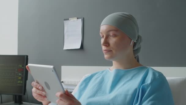 Średnie ujęcie młodej pacjentki w słuchawkach komunikujących się z przyjaciółmi na wideo przy użyciu tabletu cyfrowego leżącego w łóżku na oddziale onkologicznym - Materiał filmowy, wideo