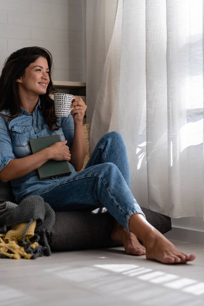 Красива усміхнена молода жінка сидить удома у затишному кутку вікна, п'є каву, розслабляється, насолоджується миттю миру - Фото, зображення