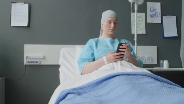 Közepes távolságból fiatal női beteg onkológiai osztály pihen az ágyon a kórteremben a modern kórház okostelefonnal - Felvétel, videó