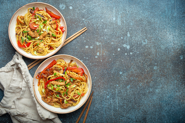 Two bowls with Chow Mein or Lo Mein, traditional Chinese stir fry noodles with meat and vegetables, served with chopsticks top view on rustic blue concrete background, space for text. - Photo, Image