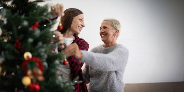 Семейный зимний отдых и концепция счастливая пожилая женщина со взрослой дочерью украшают елку дома. - Фото, изображение