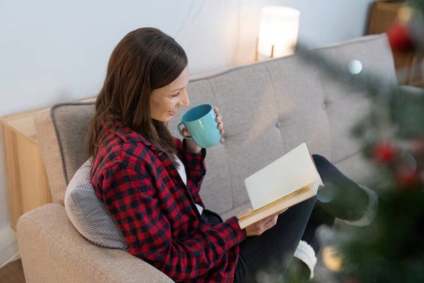 Jeune femme mignonne tenant tasse de café ou de thé et lire livre d'intérêt. Concept d'ambiance de Noël à la maison. - Photo, image