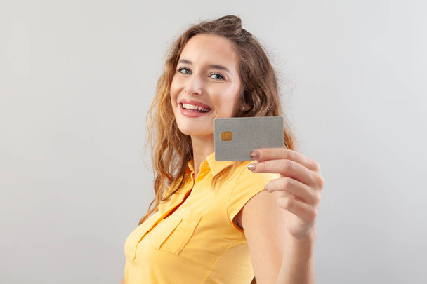 Portrait of laughing girlish woman with long hair do wear yellow top. Hand showing credit card isolated on white background - Photo, Image