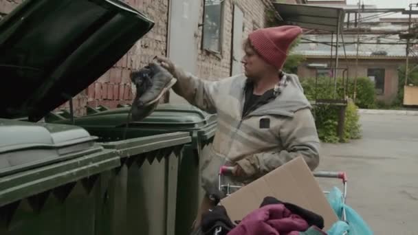Medium shot van dakloze man in lompen nemen schoenen uit prullenbak en zet ze in zijn winkelwagen, terwijl het doorbrengen van tijd buiten - Video