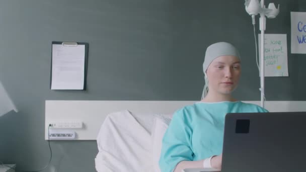 Medium shot van jonge vrouwelijke patiënt met kanker videocalling haar vrienden op laptop tijdens het zitten op bed in ziekenhuizen afdeling - Video