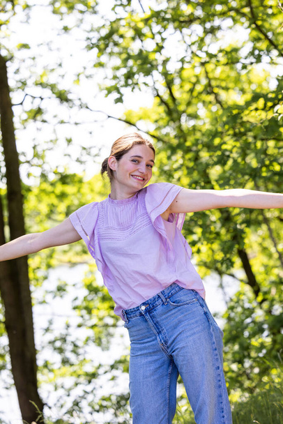 In a peaceful forest, a young woman stands side-on, her arms outstretched, embracing the warm, sunlit day. Surrounded by trees, she embodies a spirit of freedom and joy, deeply connected to the - Photo, Image
