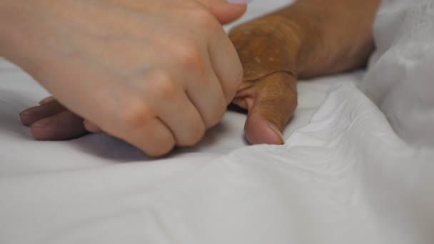 Tyttö ottaa ja varovasti koskettaa käsi sairas kypsä nainen makaa sängyssä sairaalassa. Lapsenlapsi lohduttaa vanhuksen isoäitinsä ryppyistä kättä lääketieteellisessä klinikassa. Tytär tukee äitiään. - Materiaali, video