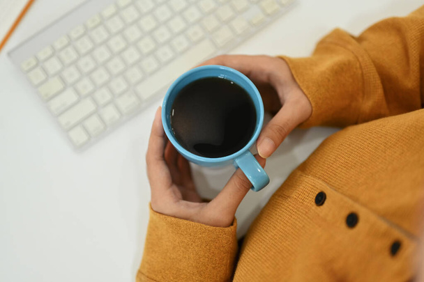 Über der Ansicht einer Frau im gelben Pullover mit einer Tasse heißen Kaffees auf dem Schreibtisch. - Foto, Bild