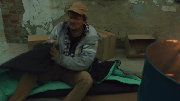 Średnie ujęcie bezdomnego zasypiającego na tekturze obok pudełek i beczki - Materiał filmowy, wideo