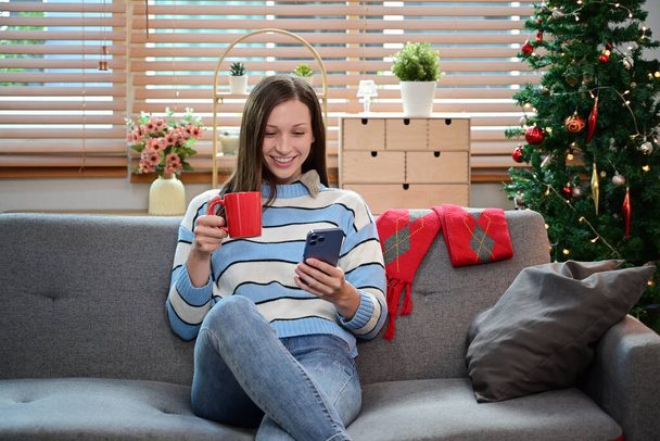 Belle femme caucasienne buvant du café et utilisant un smartphone sur un canapé près d'un arbre de Noël décoré. - Photo, image