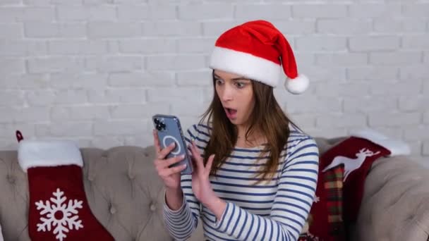 Verrassende dame met kerstmuts met smartphone, leesbericht terwijl ze thuis op de bank zit. Vrouw die naar het telefoonscherm kijkt en schokkend nieuws leest. Video in slow motion - Video