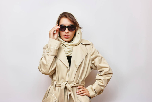 A beautiful girl in sunglasses is dressed in a light autumn raincoat and a scarf. - Photo, Image