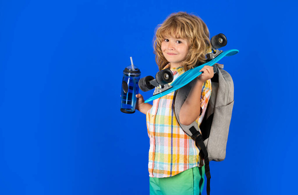 Studio ritratto di elegante carino ragazzo bambino con skateboard e bottiglia d'acqua isolato sullo sfondo blu. Pattinatore per bambini. Infanzia e tempo felice. Bambino carino con skateboard e zaino - Foto, immagini