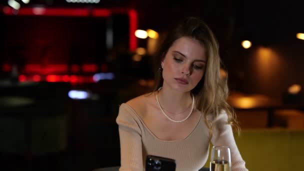 Egy fiatal hölgy pezsgőt iszik egy étteremben, és üzeneteket olvas a mobilján. Életmód koncepció. Valós idejű. - Felvétel, videó