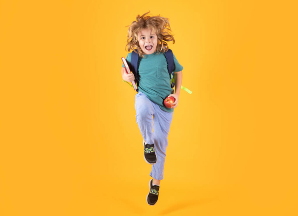 Foto von aufgeregten kleinen Schule springen mit Buch, leeren Raum isoliert gelben Hintergrund. Staunende Schulkinder springen mit Rucksack. Lernen und Wissen. Gehen Sie studieren. Bildungskonzept. Laufen und springen - Foto, Bild