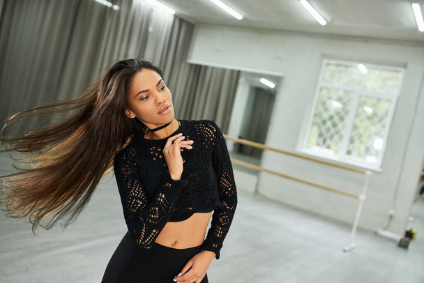 creative long-haired african american woman in black attire rehearsing rhythmic dance in modern hall - Photo, Image