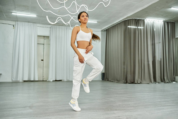 atractiva bailarina afroamericana en ropa deportiva blanca ensayando danza rítmica en estudio moderno - Foto, Imagen
