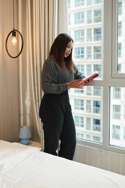 ホテルの部屋の窓に立ってプランナーをチェックするエレガントな女性起業家 - 写真・画像