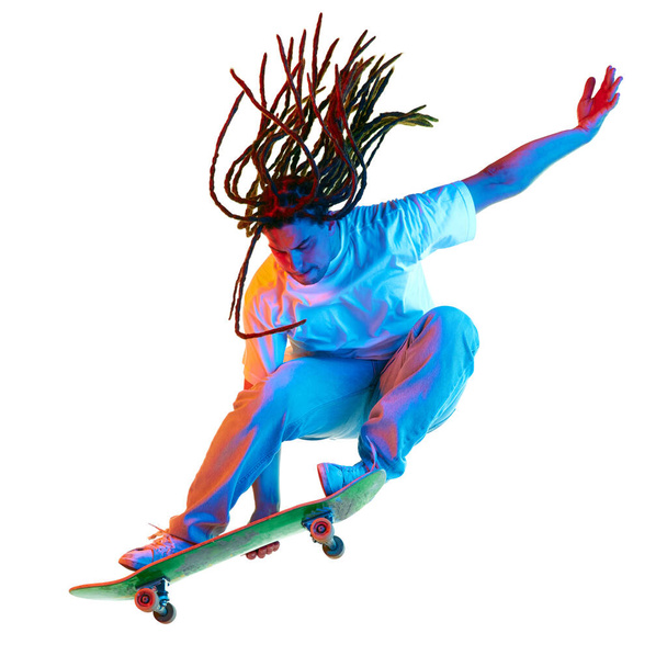 Contemporary extreme kind of sport. Man, skateboarder performing tricks in mixed neon light isolated white background. Concept of youth culture, active lifestyle, style and fashion, creativity, art. - Фото, зображення