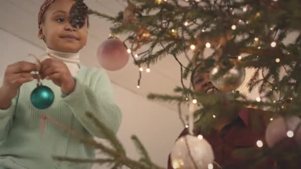 Low angle shot of African American 6 year old girl with mum decorating Christmas tree together with beautiful toys - Footage, Video