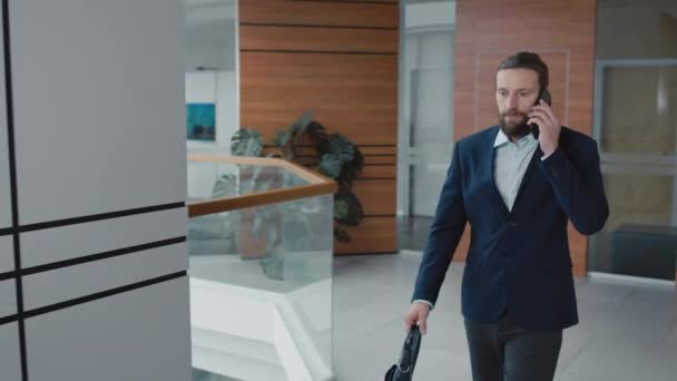 Kavkazský kancelářský pracovník v obleku mluvit na smartphone s klientem při chůzi v hale - Záběry, video